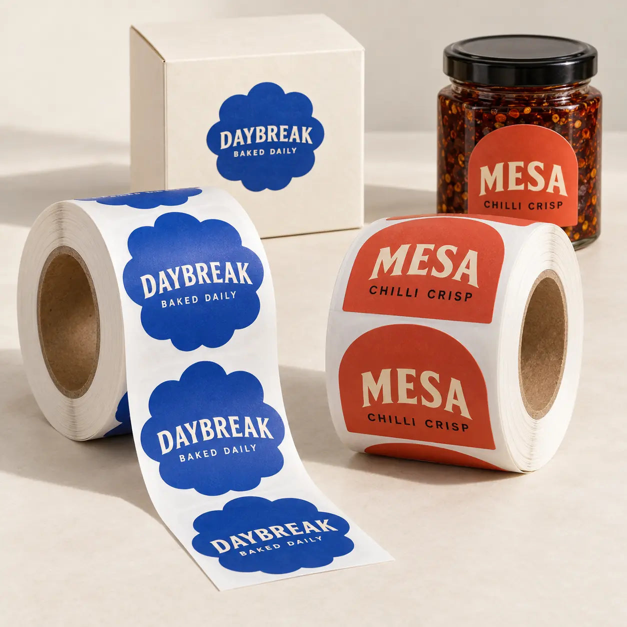 Blue DAYBREAK scalloped labels and orange MESA arch-shaped labels on two rolls, with matching labels on a bakery box and chilli crisp jar — packaging design example.